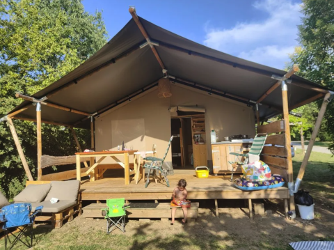 Una tenda safari con veranda, tavoli, sedie e un bambino seduto, immersa nel verde e sotto il cielo blu.