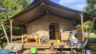 Una tienda safari con porche, mesas, sillas y un niño pequeño, rodeada de árboles y cielo azul.
