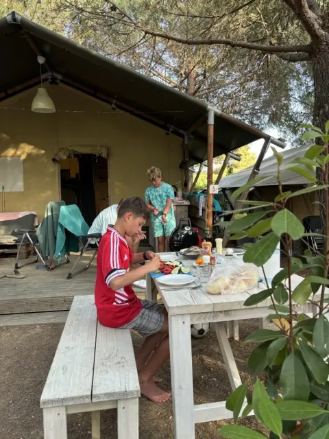Zwei Jungen essen an einem Holztisch vor einem Safarizelt, umgeben von Bäumen und Camping-Atmosphäre.