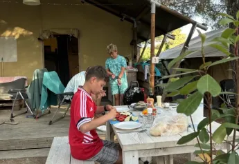 Zwei Jungen essen an einem Holztisch vor einem Safarizelt, umgeben von Bäumen und Camping-Atmosphäre.