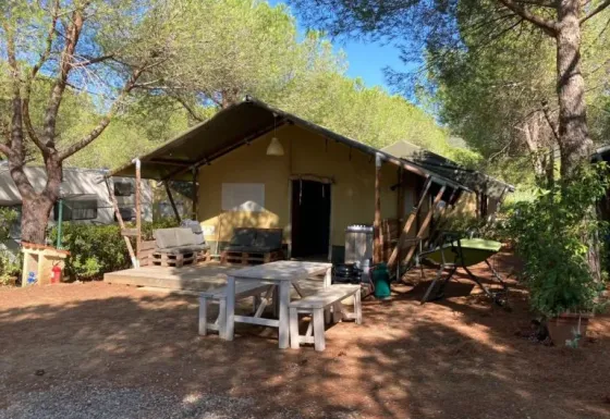 Safari-Zelt mit Holzterrasse und Sitzgelegenheiten unter Bäumen in einer bewaldeten Umgebung.