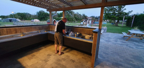 Mand vasker op i fælles udekøkken på Tornby Strand Camping med legeplads og telte i baggrunden.
