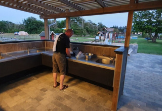 Hombre lavando platos en la cocina exterior compartida de Tornby Strand Camping, con zona de juegos y tiendas.