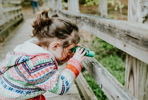Meisje in kleurrige trui kijkt door groene verrekijker vanaf houten brug in een glamping vakantiepark.