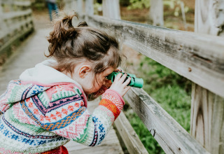 Meisje in kleurrige trui kijkt door groene verrekijker vanaf houten brug in een glamping vakantiepark.