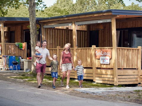 A family walks outside SunLodge Katsura at Campsite Gasparina in Italy, with wooden lodges and trees visible.
