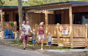 A family walks outside SunLodge Katsura at Campsite Gasparina in Italy, with wooden lodges and trees visible.