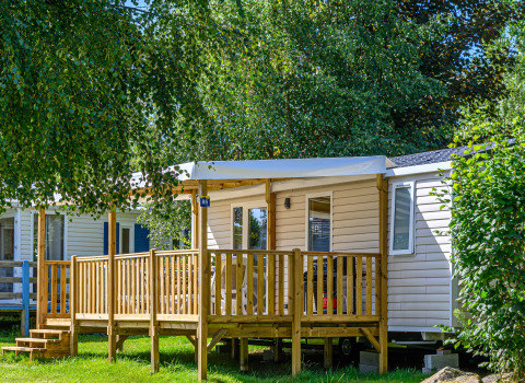 Vista exterior de Mobile home Premium con porche de madera rodeado de árboles y vegetación al sol.