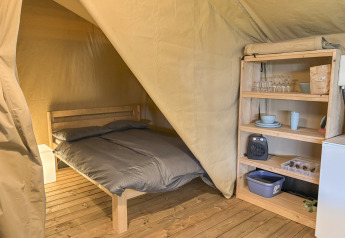 Interior de tienda safari con suelo de madera, cama doble y estantería con vajilla en Skovly Camping, Dinamarca.