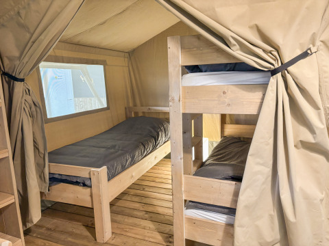 Innenaufnahme eines Safarizelts mit Einzelbett, Etagenbett, Holzboden und beigen Vorhängen.
