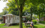 Two people and a dog sit at a picnic table outside a white cabin at Campsite Mareveld in the Netherlands.