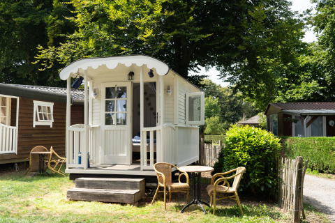 Casetta Pipowagen bianca con veranda e sedie da giardino, immersa nel verde tra alberi e siepi.