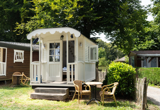 Casetta Pipowagen bianca con veranda e sedie da giardino, immersa nel verde tra alberi e siepi.