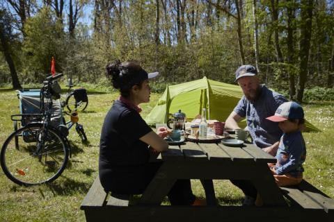Gezin picknickt aan een houten tafel met tent en fietsen bij Nordskoven Strand Camping, Denemarken.