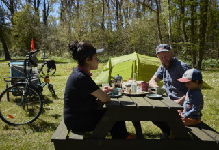 Gezin picknickt aan een houten tafel met tent en fietsen bij Nordskoven Strand Camping, Denemarken.