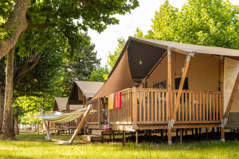 Safari tent called Villatent Outback with a wooden deck and hammock, set among green trees on a sunny day.