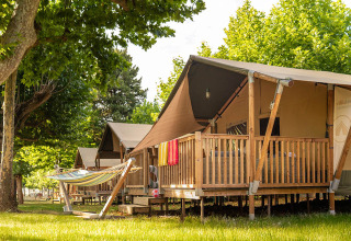 Safari tent genaamd Villatent Outback met houten veranda en hangmat onder groene bomen op een zonnige dag.