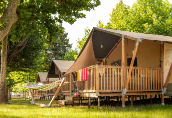 Safari tent called Villatent Outback with a wooden deck and hammock, set among green trees on a sunny day.
