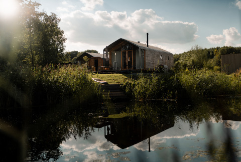 Wikkelhouse met Ligbad ved en stille dam, omgivet af natur og træer under en solrig himmel med skyer.