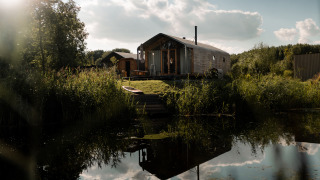 Wikkelhouse met Ligbad au bord d’un étang paisible, entouré de nature, d’arbres et d’un ciel partiellement nuageux.