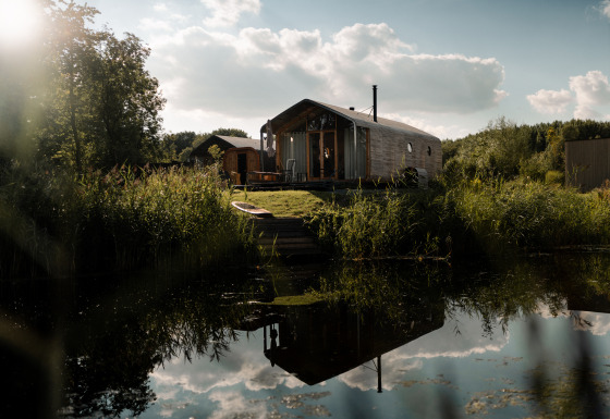 Wikkelhouse met Ligbad ved en stille dam, omgivet af natur og træer under en solrig himmel med skyer.