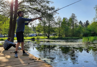 Dos personas pescando junto al estanque en Skovly Camping, rodeados de verde y tiendas en Región de Zelanda, Dinamarca.