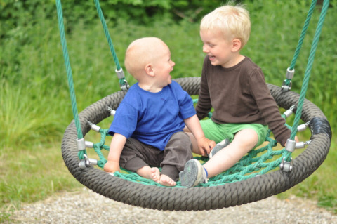 To små børn leger og smiler sammen på en rund gynge i Skovly Camping, Region Sjælland, Danmark.