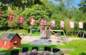 Spielplatz mit dänischen Flaggen, Feuerstelle und Holzhäuschen im Skovly Camping, Region Seeland, Dänemark.