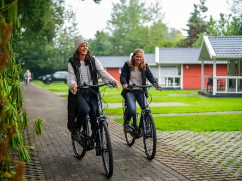 Due donne felici vanno in bicicletta nel parco vacanze Latour nel Brabante Settentrionale, Paesi Bassi, vicino ai bungalow.