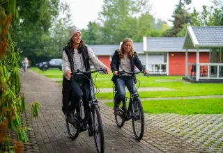 Two happy women riding bicycles on a path at Holiday park Latour in North-Brabant, Netherlands, near cabins.