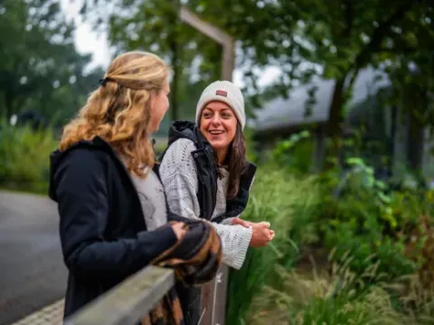 Two women chat and smile outdoors at Holiday Park Latour in North Brabant, Netherlands, among greenery.