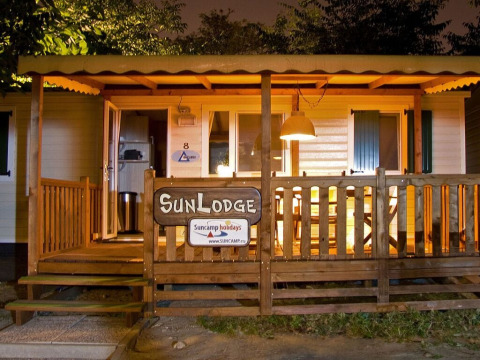 Gezellig verlichte SunLodge chalet met houten veranda en bord, perfect voor een ontspannen vakantieavond.