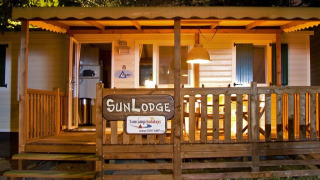 Oplyst SunLodge-hytte med træveranda og skilt, ideel til afslappende ferier i naturen om aftenen.