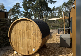 Udendørs glampingområde med en rund trætønde, hytte, træterrasse og store træer i baggrunden.