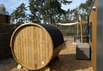 Area glamping all'aperto con sauna di legno a forma di botte, cabina moderna e alberi circostanti.