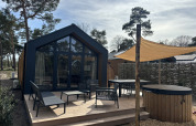 Modern Boslodge tiny house with patio, outdoor seating, sunshade, hot tub and sauna, set in a forested area.