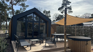 Modern Boslodge tiny house with patio, outdoor seating, sunshade, hot tub and sauna, set in a forested area.