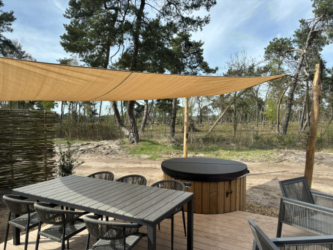 Terrasse extérieure avec table, chaises, voile d'ombrage et jacuzzi sous abri près de la forêt.