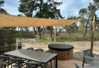 Terrasse extérieure avec table, chaises, voile d'ombrage et jacuzzi sous abri près de la forêt.