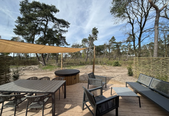 Terrazza in legno presso Boslodge con tavolo, sedie lounge, vasca idromassaggio e vista sul bosco.