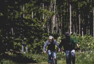 Deux personnes font du vélo sur un sentier forestier ensoleillé près d'un site de glamping, entourés d'arbres.