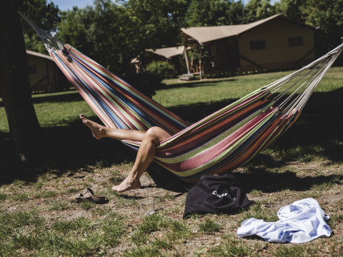 Une personne se détend dans un hamac coloré devant des tentes safari, avec chaussures et vêtement sur l’herbe.