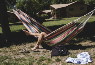 Une personne se détend dans un hamac coloré devant des tentes safari, avec chaussures et vêtement sur l’herbe.