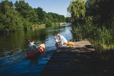 Famille en glamping au bord d’un lac : homme en kayak, femme et enfant assis sur un ponton en bois.