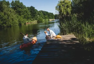 Famille en glamping au bord d’un lac : homme en kayak, femme et enfant assis sur un ponton en bois.