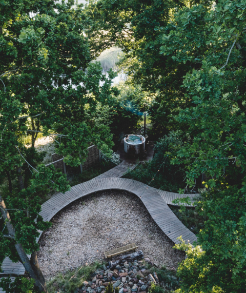 Vue aérienne d’un site de glamping avec sentier en bois, espace gravier et bain extérieur entouré d’arbres.