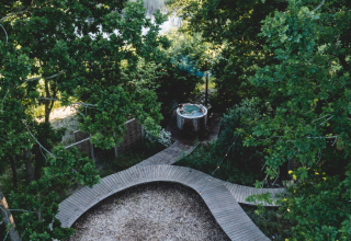 Vue aérienne d’un site de glamping avec sentier en bois, espace gravier et bain extérieur entouré d’arbres.