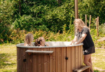 Een vrouw praat met twee kinderen die ontspannen in een hot tub bij een glamping accommodatie in de natuur.