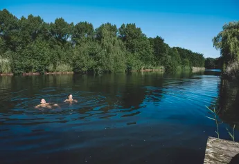 Zwei Personen schwimmen in einem ruhigen See, umgeben von grünen Bäumen und blauem Himmel beim Glamping.