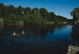 Deux personnes nagent dans un lac paisible entouré d’arbres verts sous un ciel bleu au glamping.